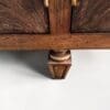 Close-up of a dark wood furniture corner with a central vertical post, brass hinges, and a carved decorative foot below, against a light background.