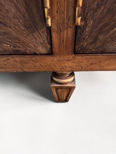 Close-up of a dark wood furniture corner with a central vertical post, brass hinges, and a carved decorative foot below, against a light background.