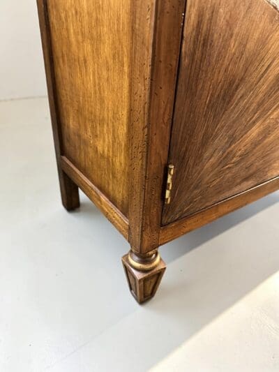 Close-up of a wooden cabinet leg with a turned, gold-accented foot and brass hardware, highlighting the wood grain.
