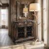 Distressed wooden cabinet with ornate iron grille, topped by a white lampshade and antler-inspired stand, in a sunlit wooden room.