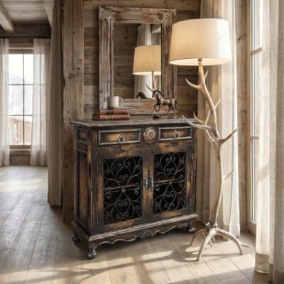 Distressed wooden cabinet with ornate iron grille, topped by a white lampshade and antler-inspired stand, in a sunlit wooden room.